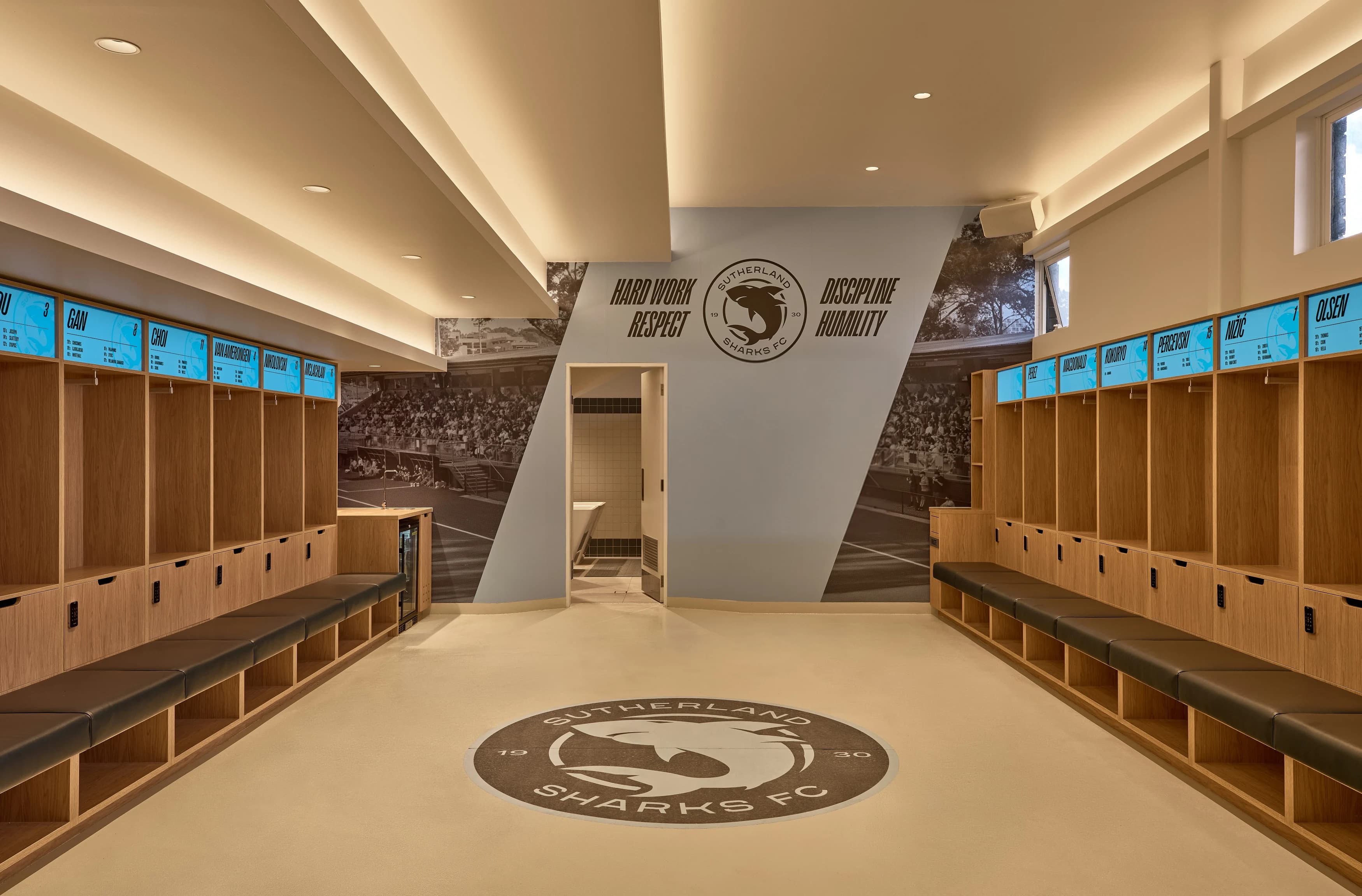 The main change room at Seymour Shaw: timber lockers under sky-blue name panels on both walls, leather benches, and the Sutherland Sharks FC crest set into the floor