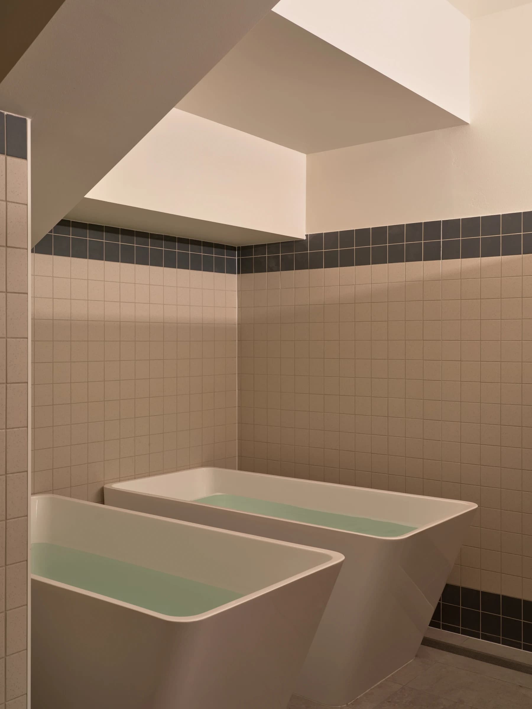 Two ice baths side by side in a tiled recovery room, lit from a skylight above