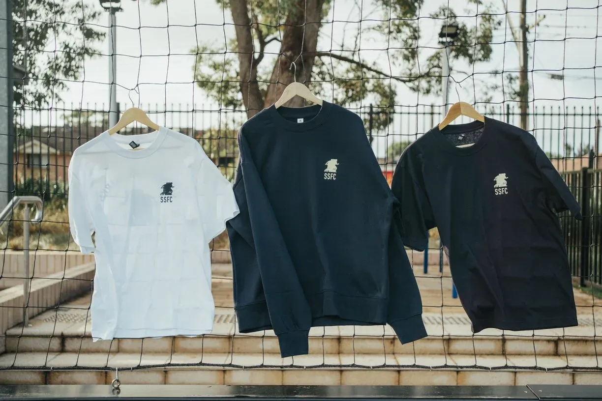 Three Sharks tops on hangers against a net: a white T-shirt, a navy sweatshirt and a navy T-shirt, each with the SSFC shark on the chest