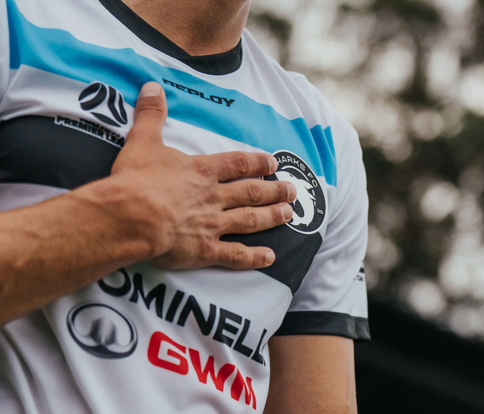 A hand on the club crest of a white Sharks shirt with Dominelli GWM on the front