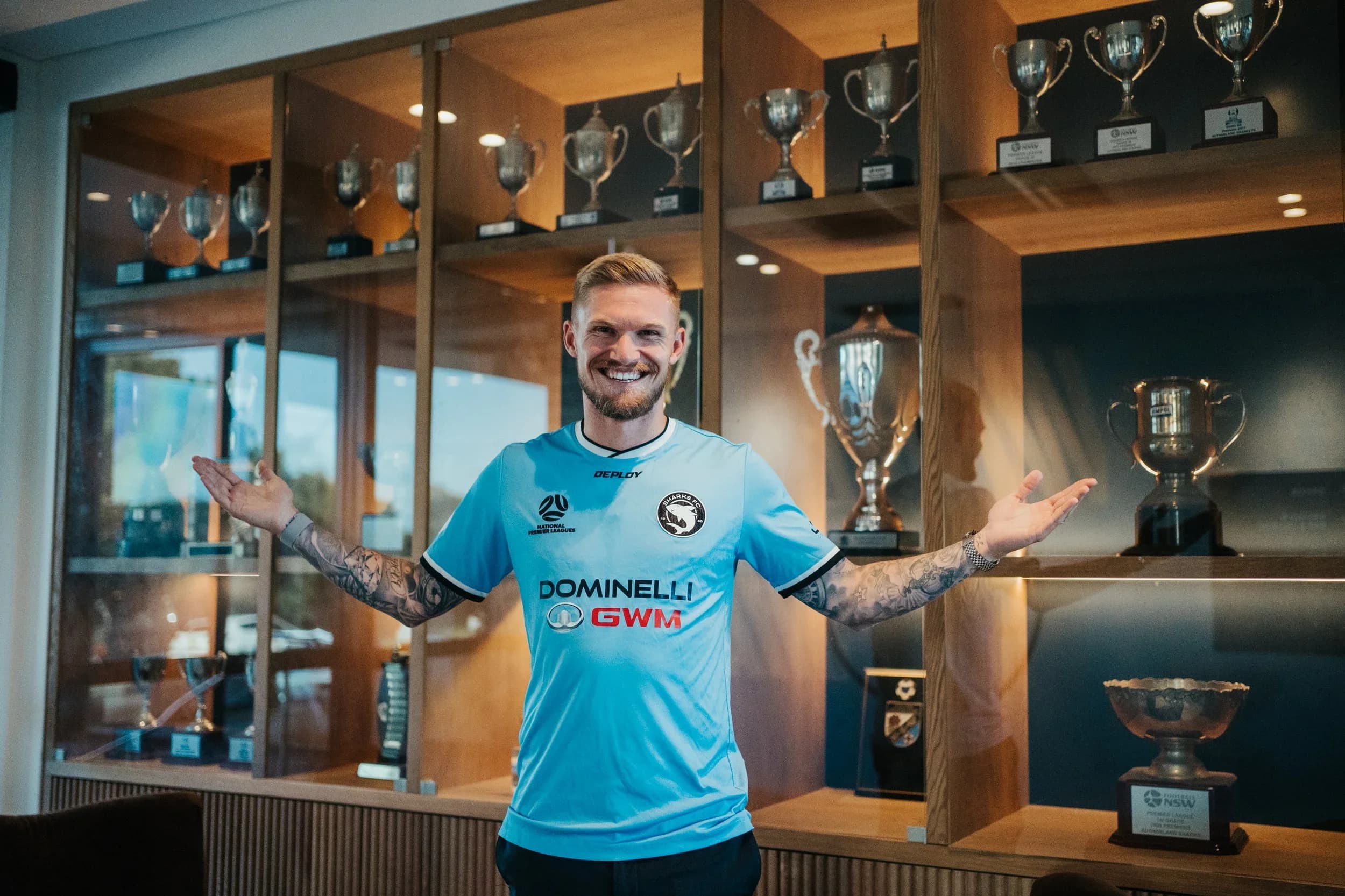 Nicholas Olsen, arms spread, in a Sharks shirt in front of the club's trophy cabinet