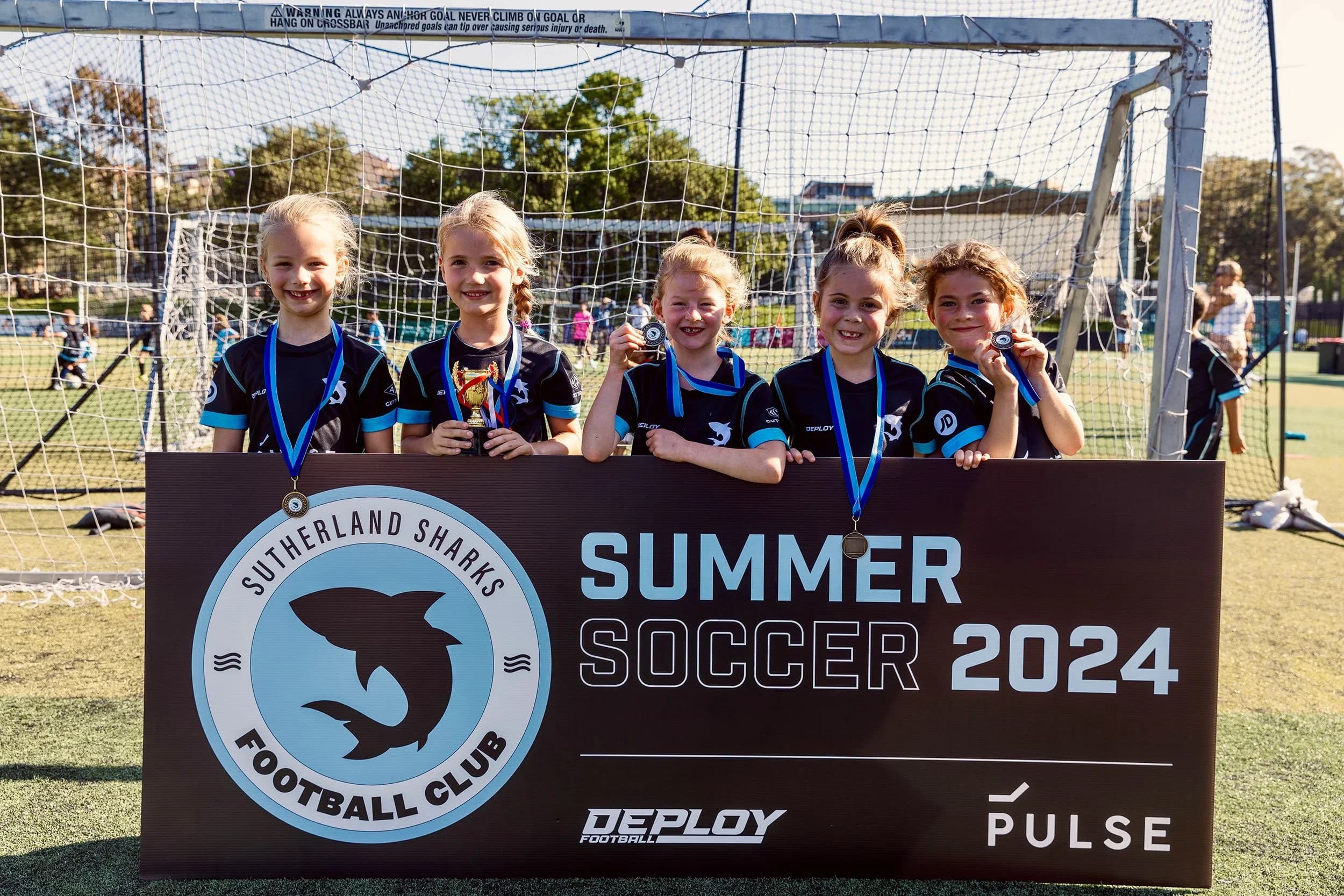 Smiling girls with medals behind the Summer Soccer 2024 banner