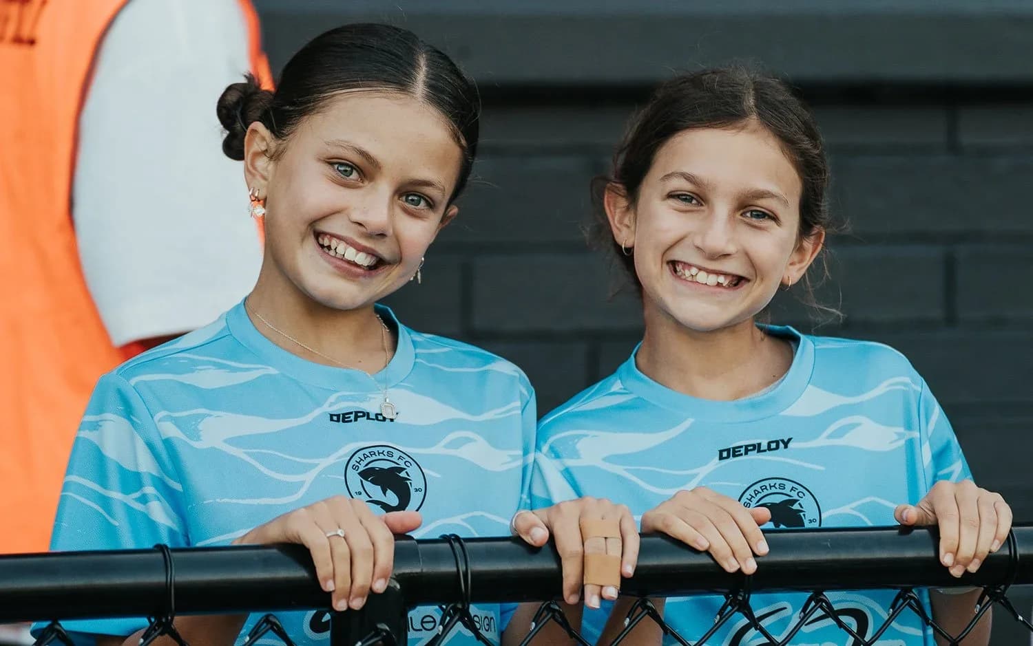 Two smiling girls in sky-blue Sharks tops lean on a fence