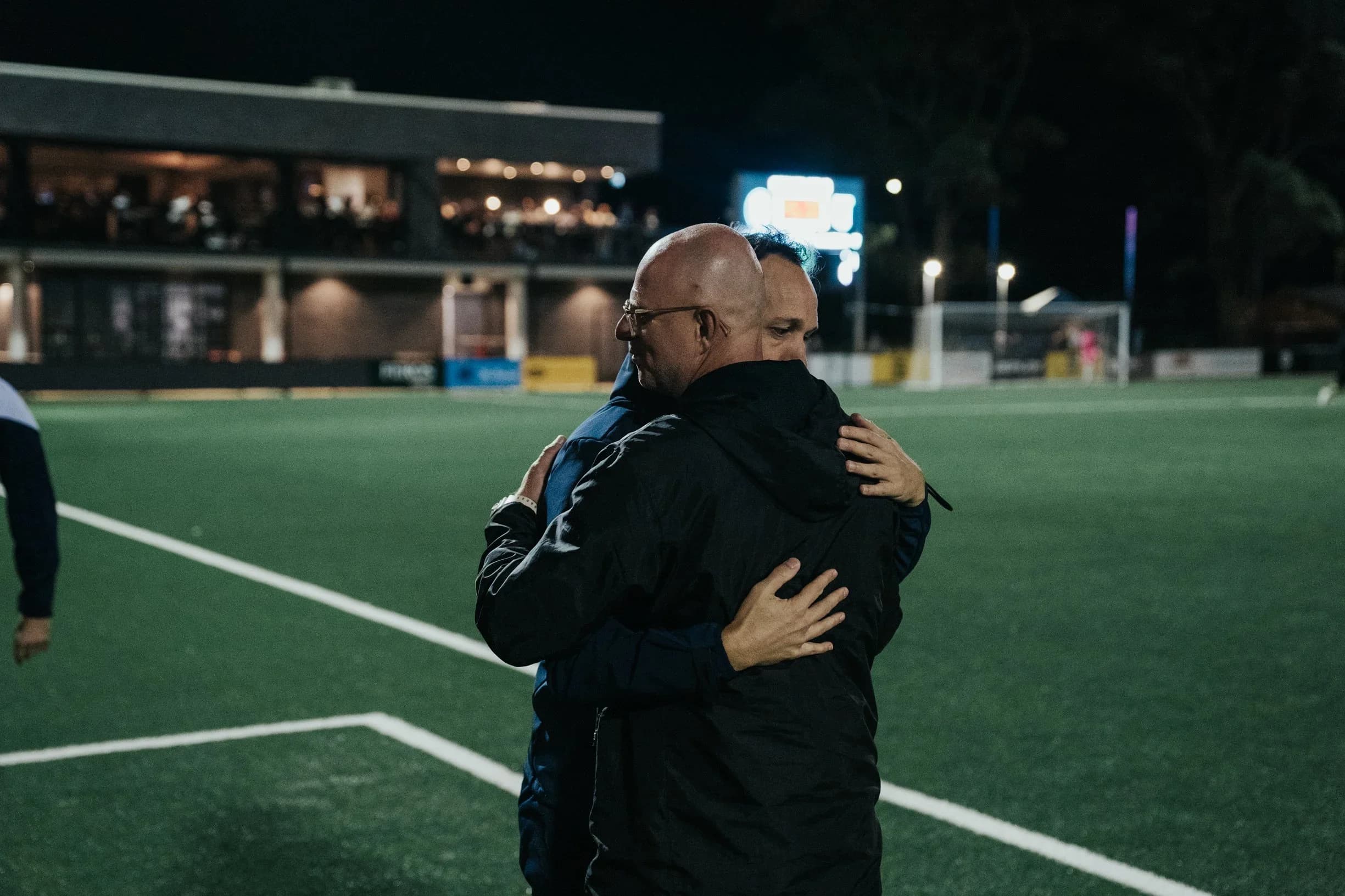 Two coaches embrace on the pitch at night after the match