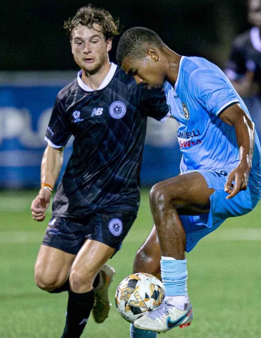 A Sharks player in sky blue and an Olympic player in navy battle for the ball at night