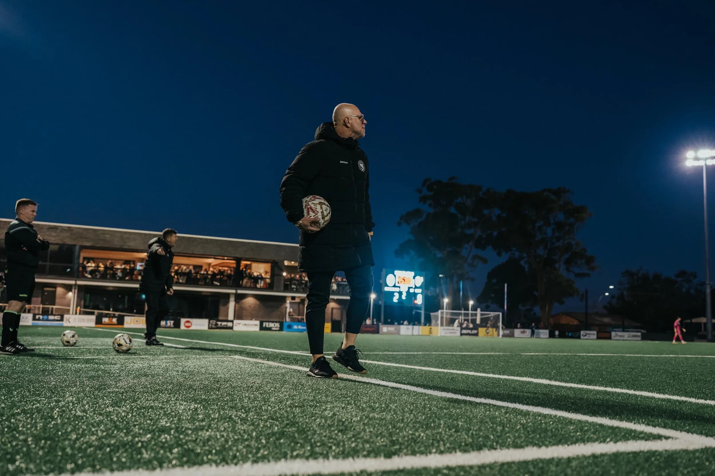 A coach walks across the pitch at night with the clubhouse lit up behind