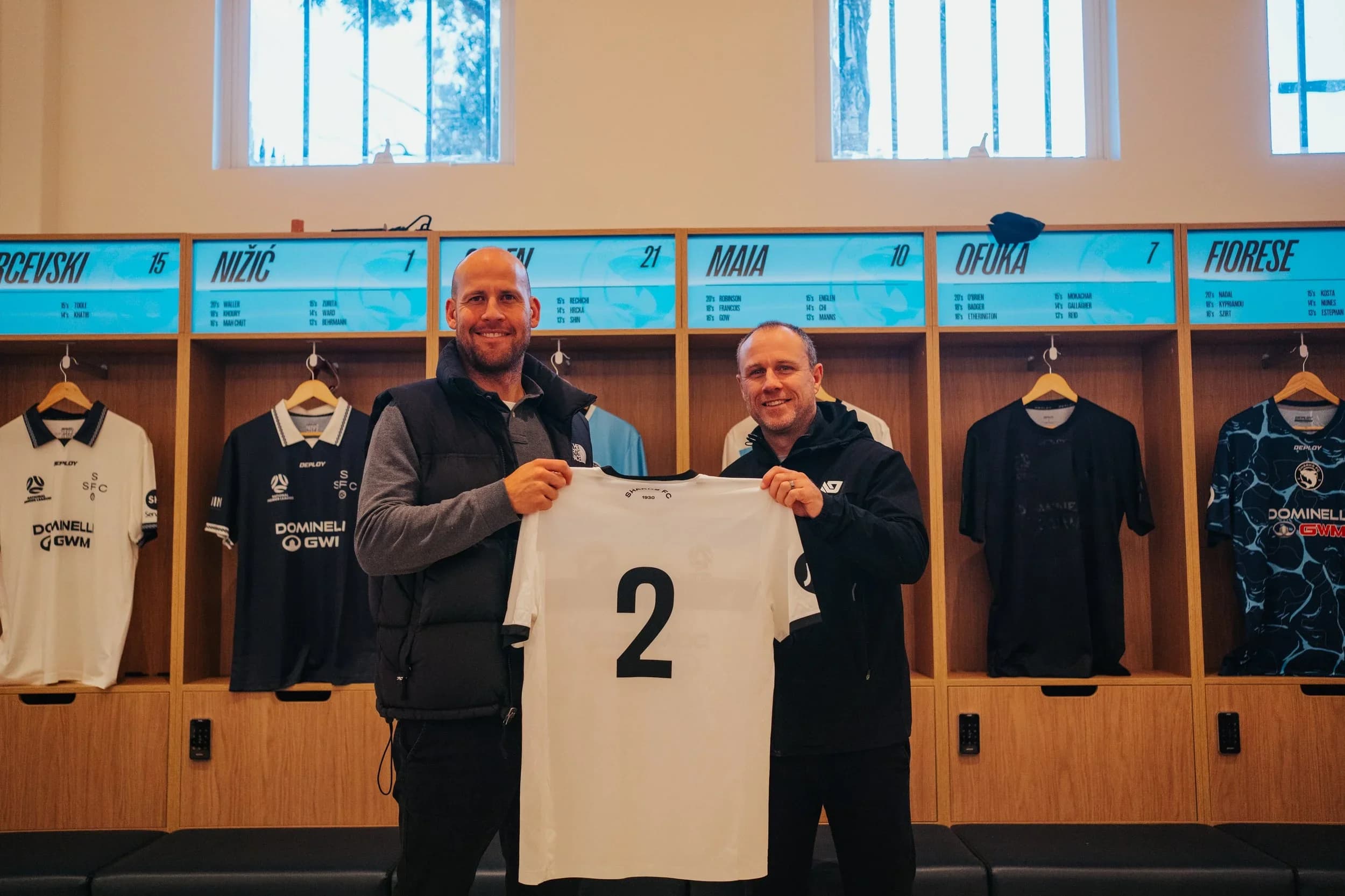 Two Deploy representatives hold up a white Sharks shirt with the number 2 in the dressing room