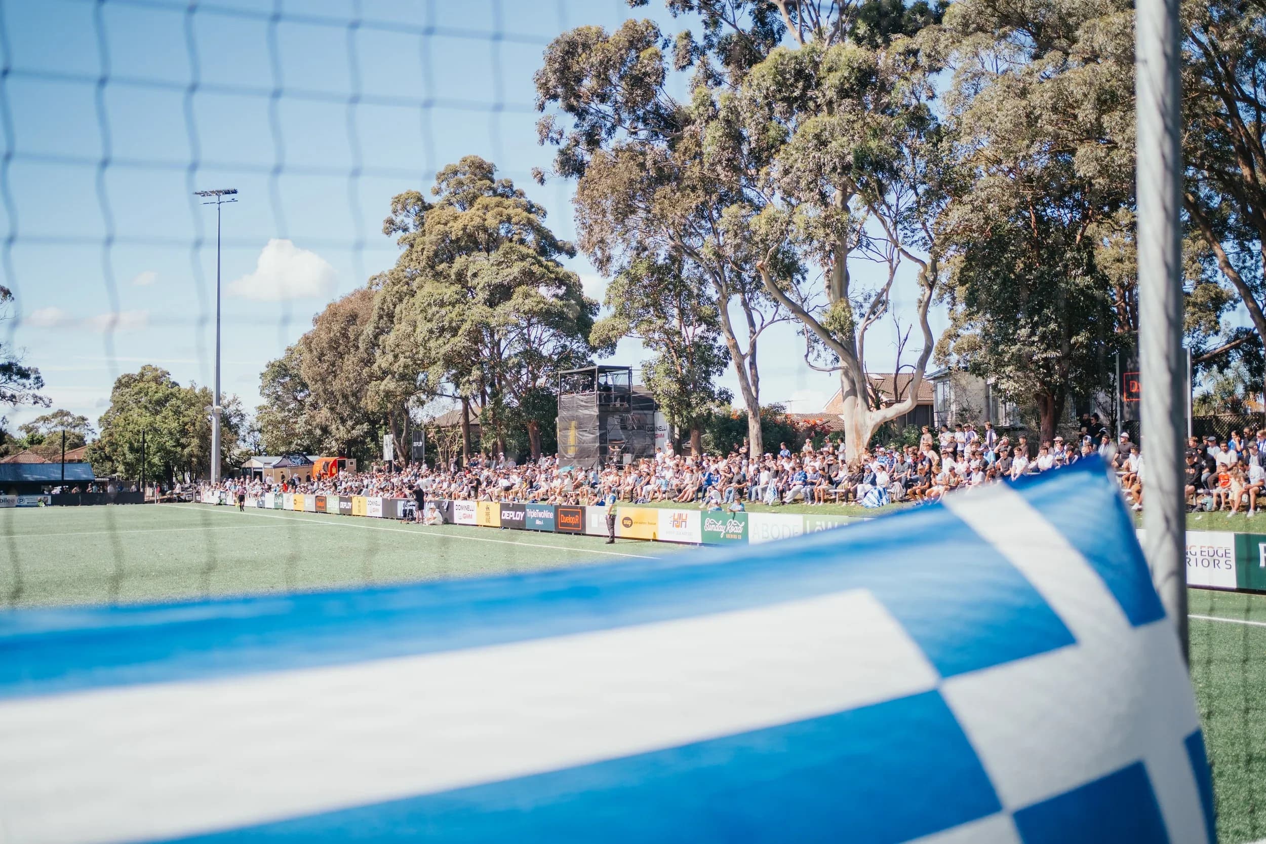 A blue and white flag in the foreground and a full crowd along the far side of the pitch
