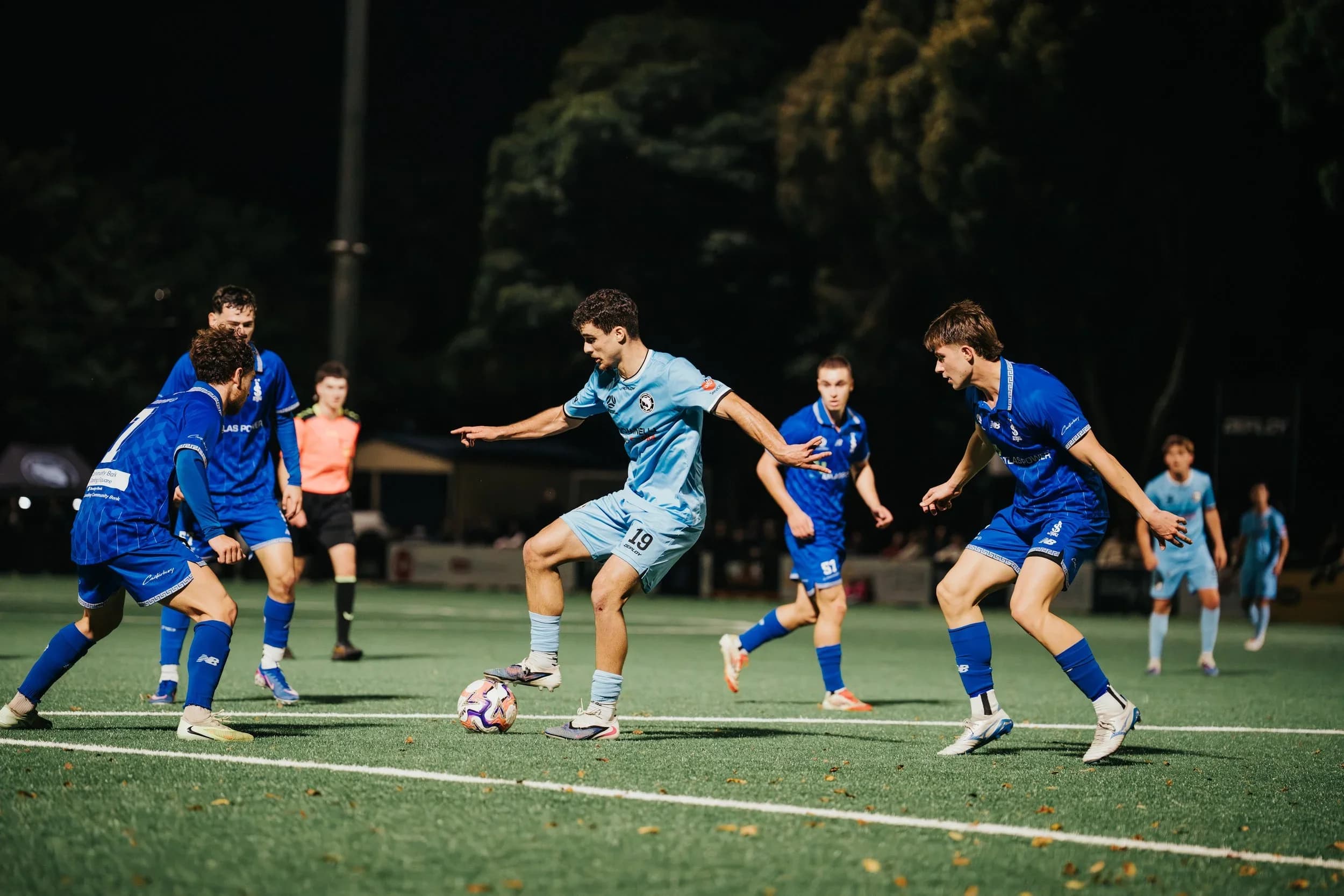 A Sharks player in sky blue runs with the ball between two opponents in blue at night