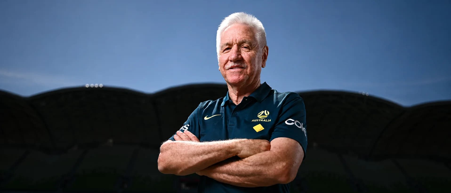 Tom Sermanni, arms folded, in a navy polo against a dusk sky