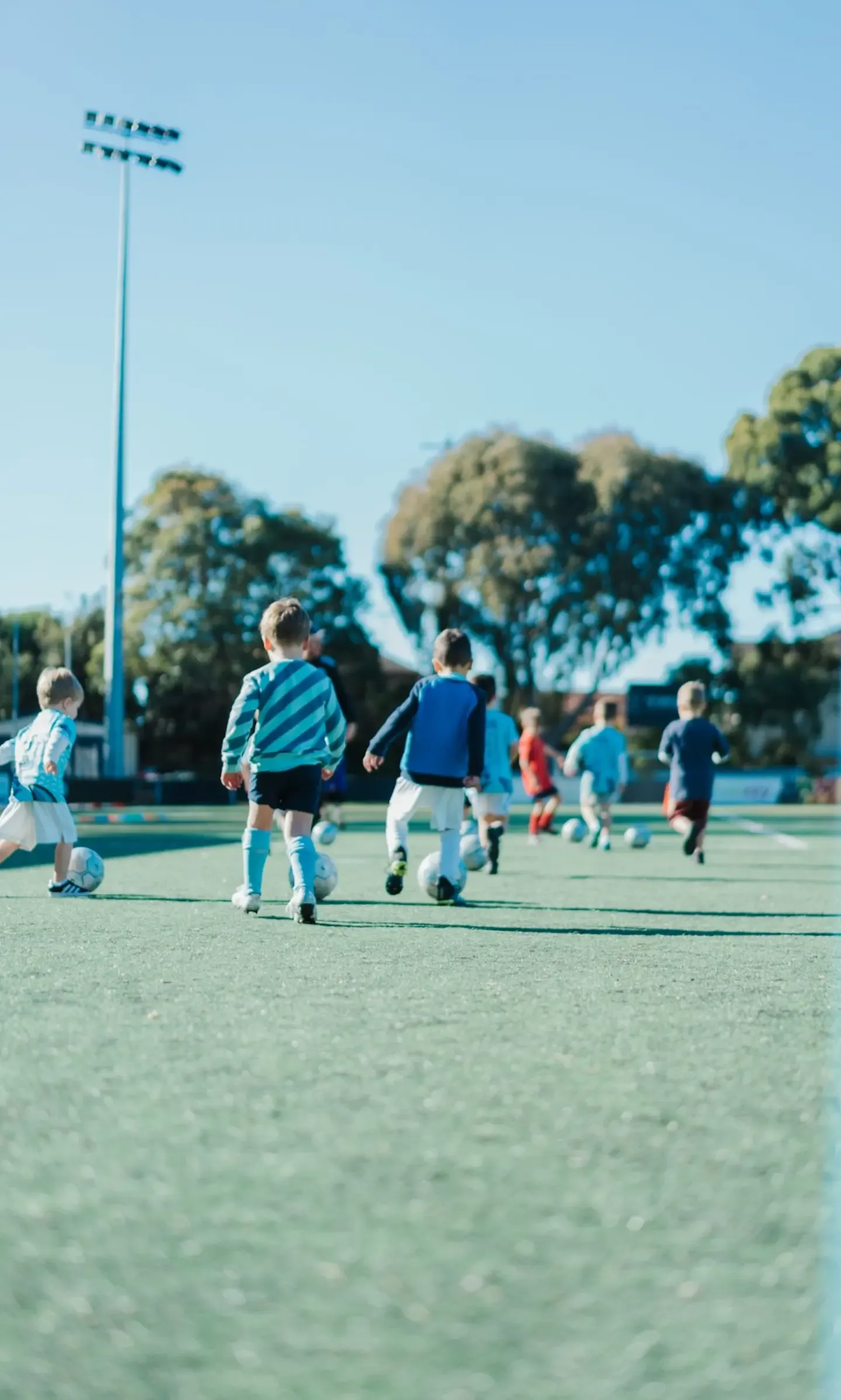 Preschoolers dribbling footballs across the pitch at Seymour Shaw on a sunny morning, seen from behind