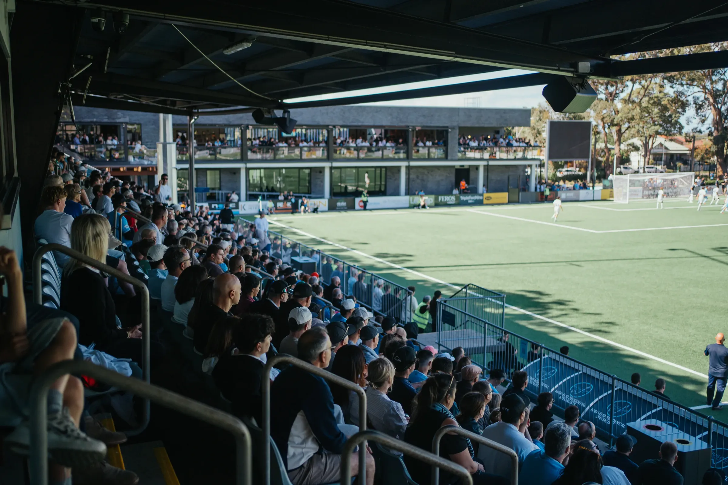 A packed stand at Seymour Shaw on a sunny afternoon, looking across the pitch to the clubhouse and its balcony full of fans