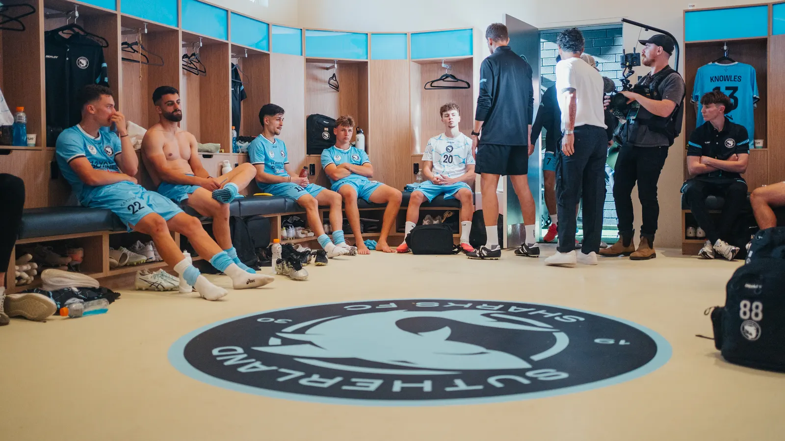 A camera operator filming in the Sharks change room on the day of the South Melbourne game, players on the benches around the crest on the floor